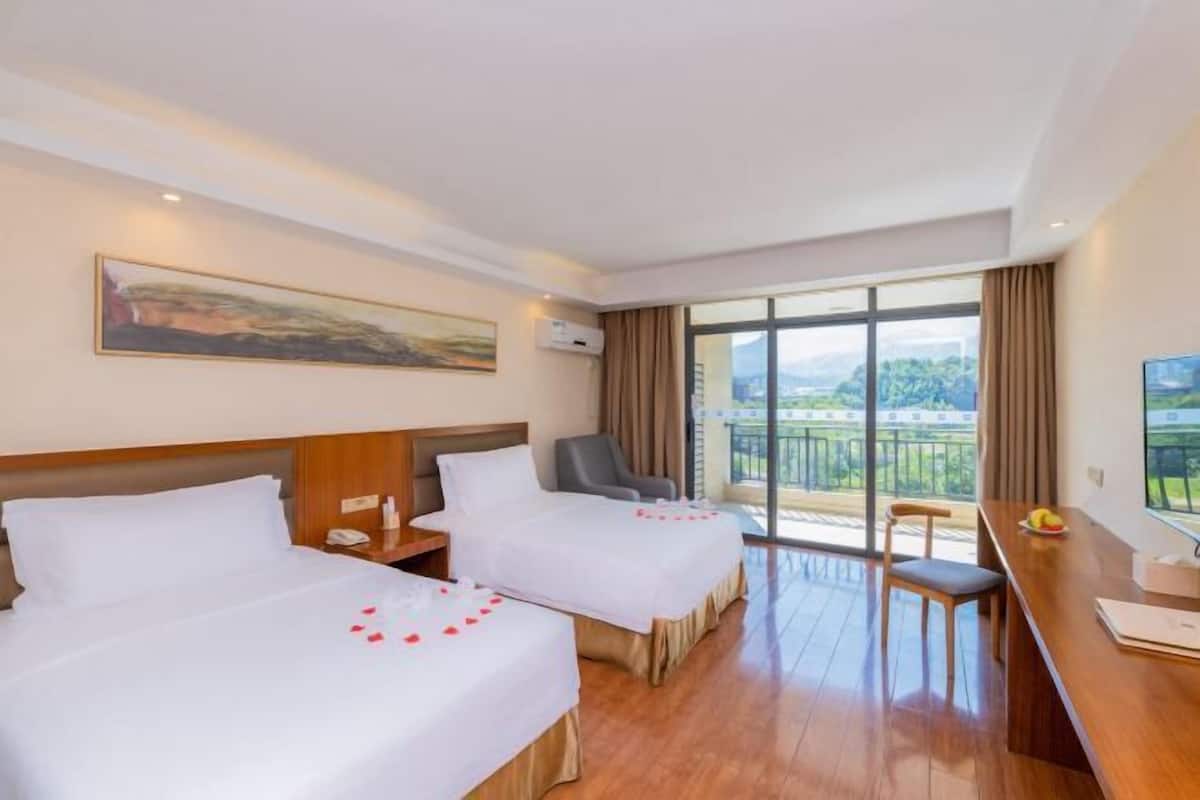 A hotel room with two beds, a desk, a chair, a painting, and a balcony with a view.