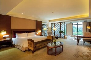 A hotel room with a large bed, a wooden armchair, a small round table, and a view of greenery through large windows.