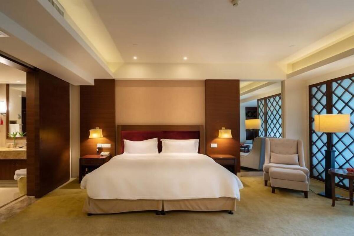 A hotel room with a large bed, bedside lamps, a chair, and a view through a lattice window.