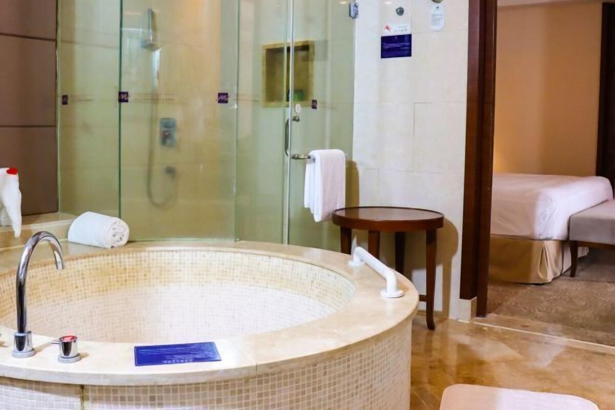 A bathroom with a glass-enclosed shower, a freestanding bathtub, a small table with a towel, and a bed with a white cover.