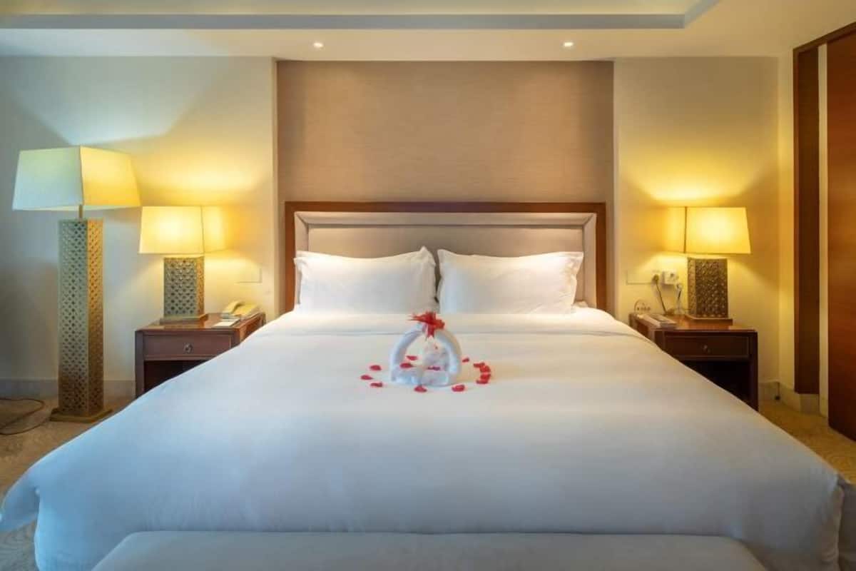 A hotel room with a large bed, two bedside tables with lamps, a headboard, and a decorative arrangement of flowers and a heart on the bed.