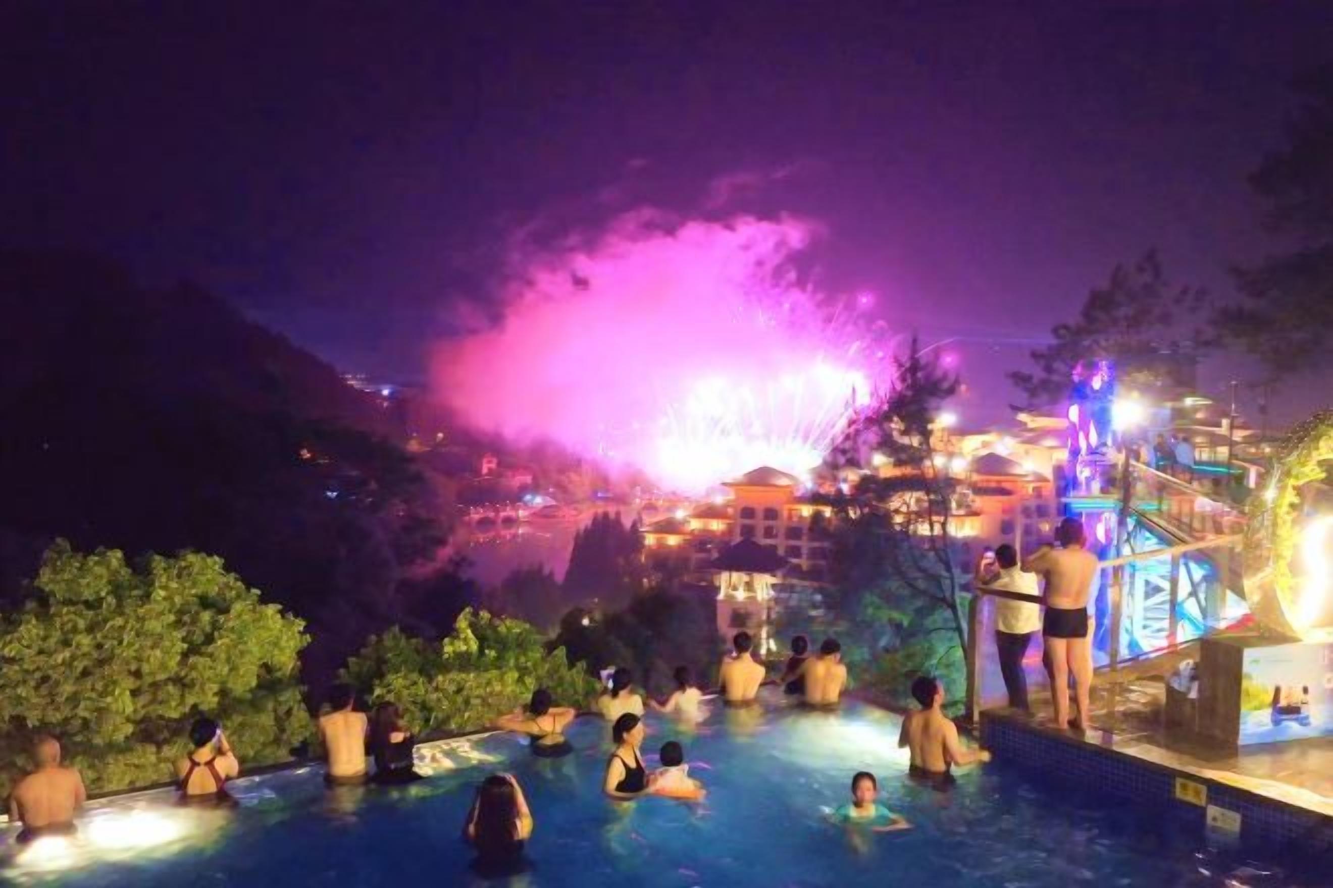 A nighttime swimming pool area with people relaxing, a water fountain display, and a brightly lit building in the background.