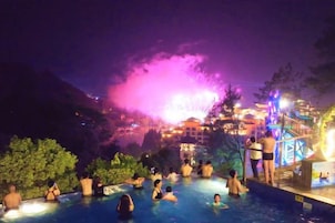 Zona de piscina nocturna con gente relajándose, un espectáculo de fuente de agua y un edificio iluminado al fondo.