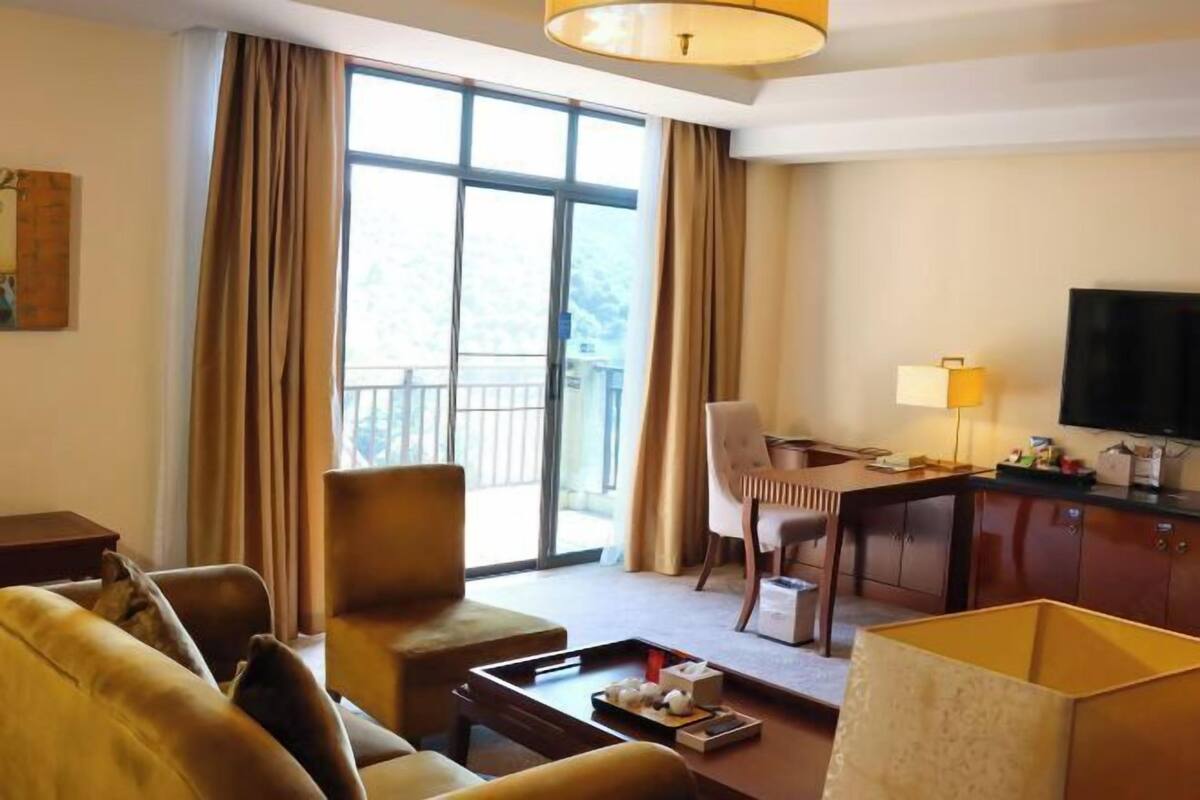 A hotel room with a sofa, a dining table, a TV, and a balcony with curtains.