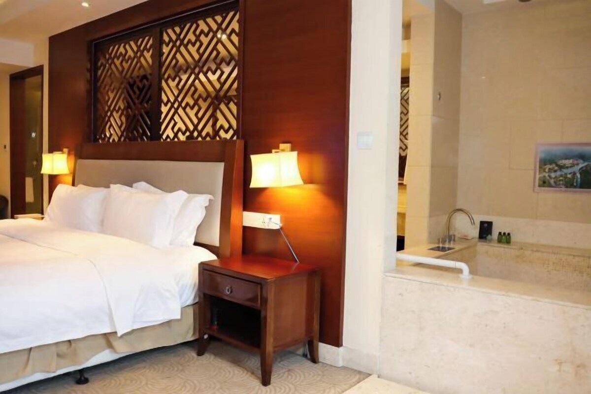 A hotel room with a large bed, two bedside lamps, a wooden headboard, a small nightstand, a bathroom with a bathtub, and a tiled wall.