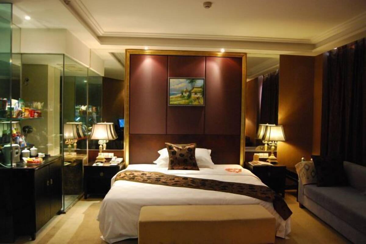 A hotel room with a large bed, bedside tables with lamps, a glass partition, and a painting on the wall.