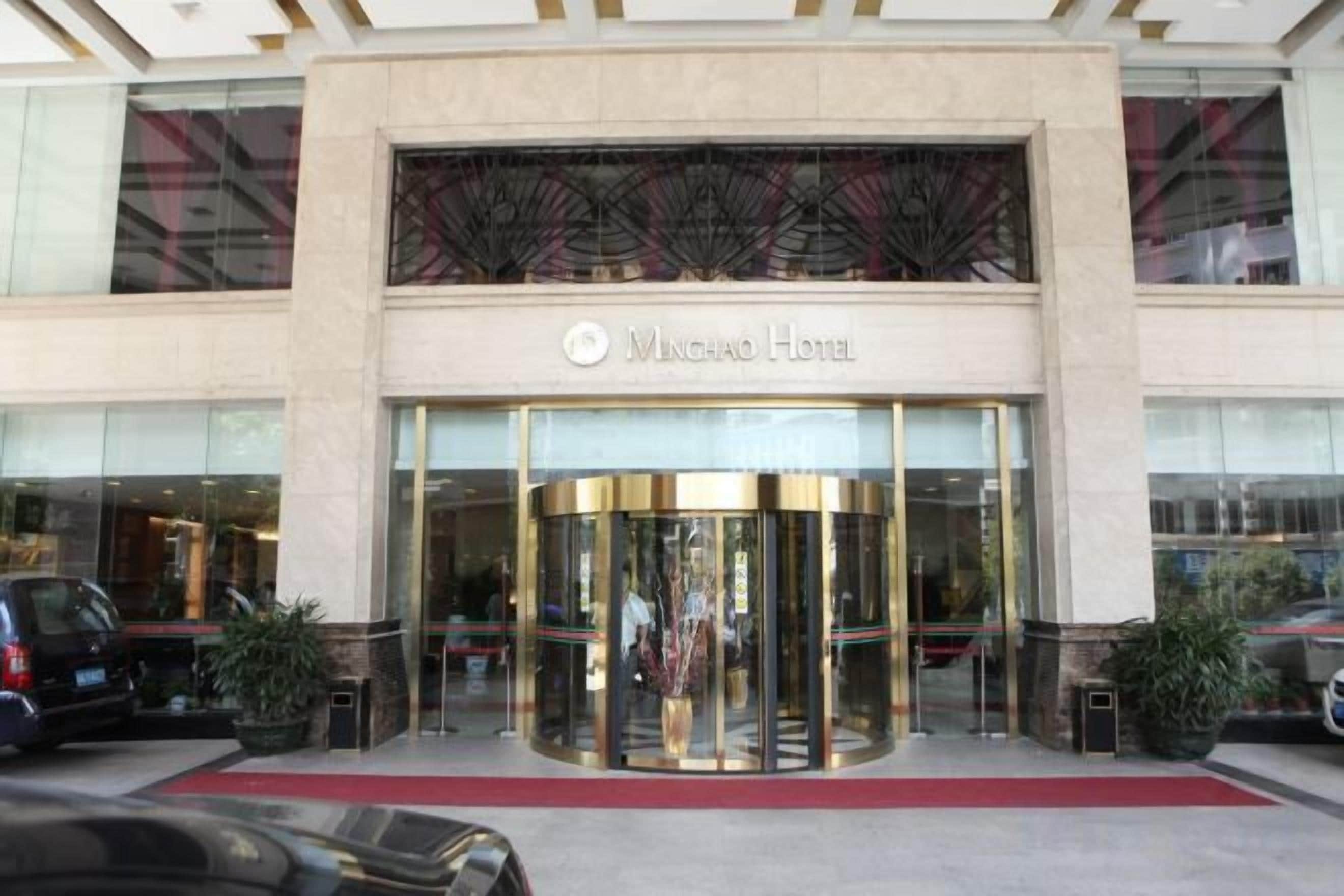 The exterior of the Minghao Hotel with a revolving door entrance, large glass windows, and a red carpet leading to the entrance.