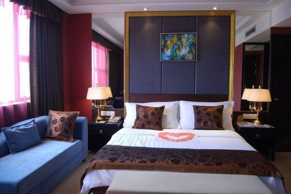 A hotel room with a bed, a sofa, two bedside tables with lamps, a painting on the wall, and a heart-shaped decoration on the bed.