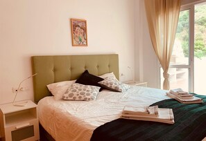 A neatly made bed with a green headboard, white bedding, patterned pillows, and a view of greenery through a window.