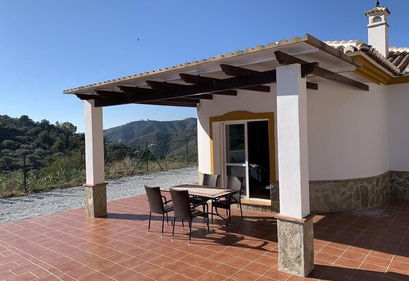 A covered patio with a table and chairs, a view of hills, and a building with a tiled roof.
