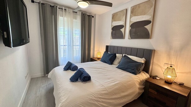 A hotel room with a bed, a nightstand, a lamp, a ceiling fan, and artwork on the wall.