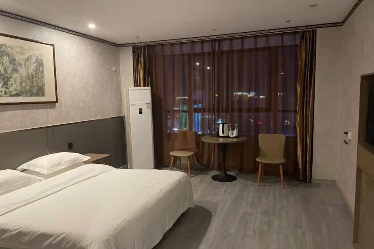 A hotel room with a bed, a small table with chairs, a refrigerator, and a large window with curtains.