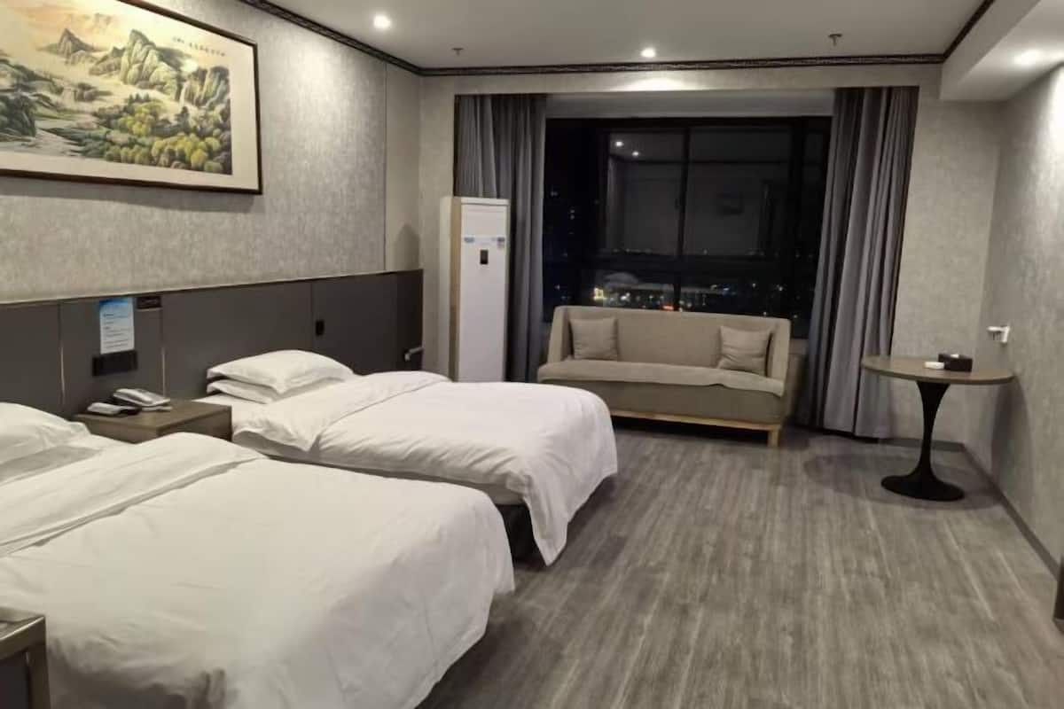 A hotel room with two beds, a sofa, a small table, a painting on the wall, and a view of the city at night.