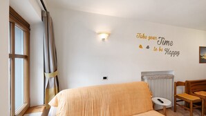 Une pièce comprenant un lit, une chaise, une petite table, un luminaire fixé au mur, un radiateur et une fenêtre avec des rideaux.