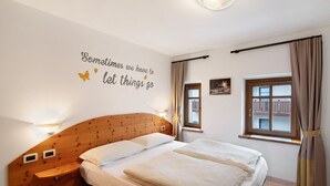Une chambre avec une tête de lit en bois, un lit avec une literie blanche, une lampe fixée au mur, une fenêtre avec des rideaux et une citation inspirante sur le mur.