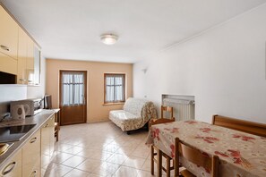 Quarto pequeno e bem iluminado com cozinha compacta, uma cama, uma cadeira e duas janelas.