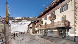 Une rue enneigée, avec un bar, un magasin appelé « Chaussures et Sacs », et une montagne en arrière-plan.