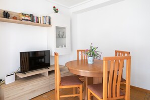 A dining area with a round table, four wooden chairs, a television on a stand, a bookshelf, and a small potted plant.