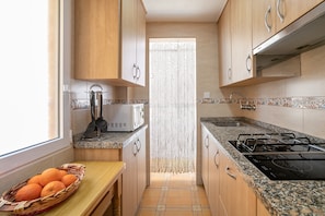 A narrow kitchen with wooden cabinets, a microwave, a gas stove, and a window with a basket of oranges.
