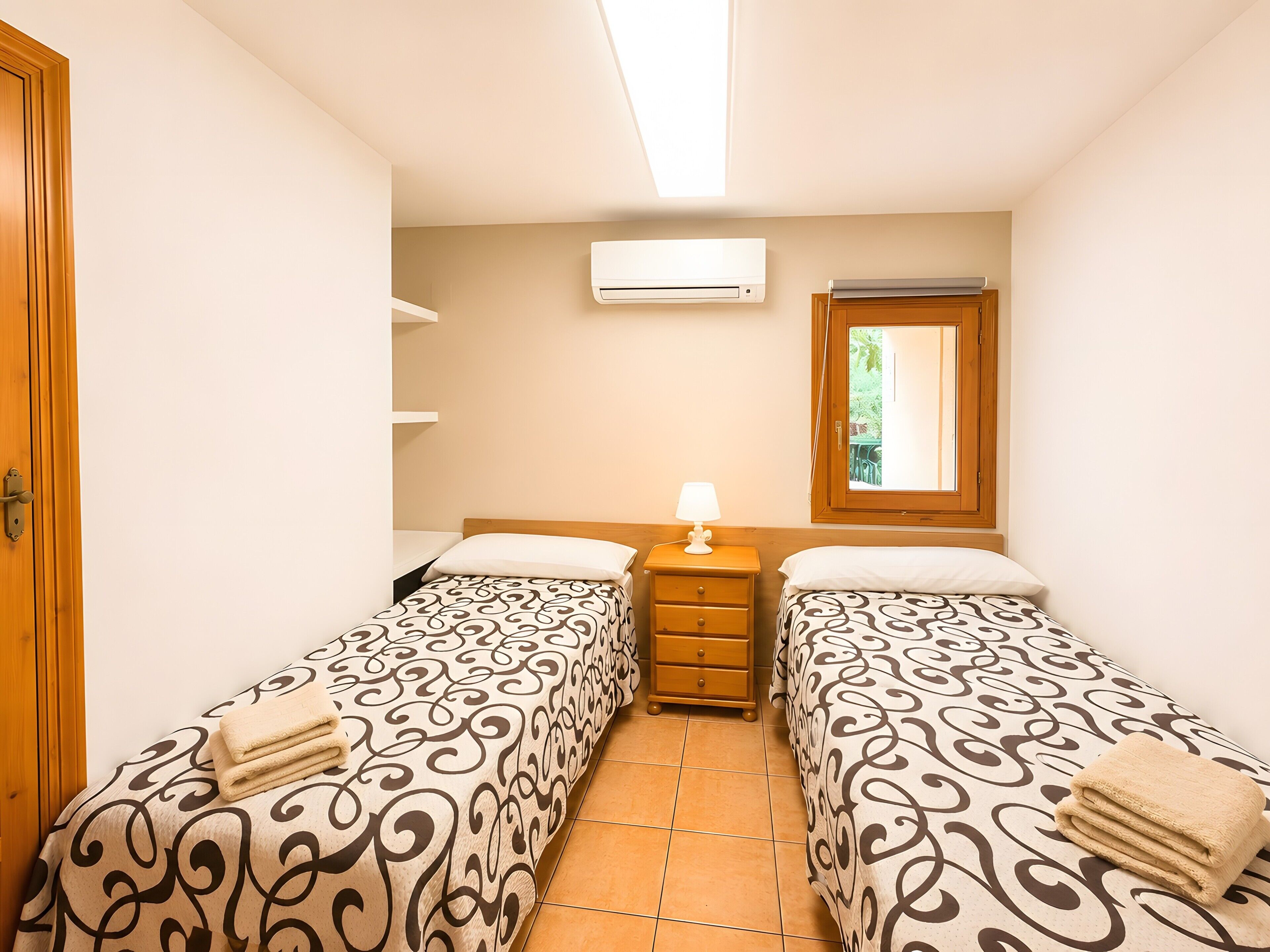 Une chambre avec deux lits simples, une table de chevet avec une lampe, un appareil de climatisation et une fenêtre donnant sur un paysage verdoyant.