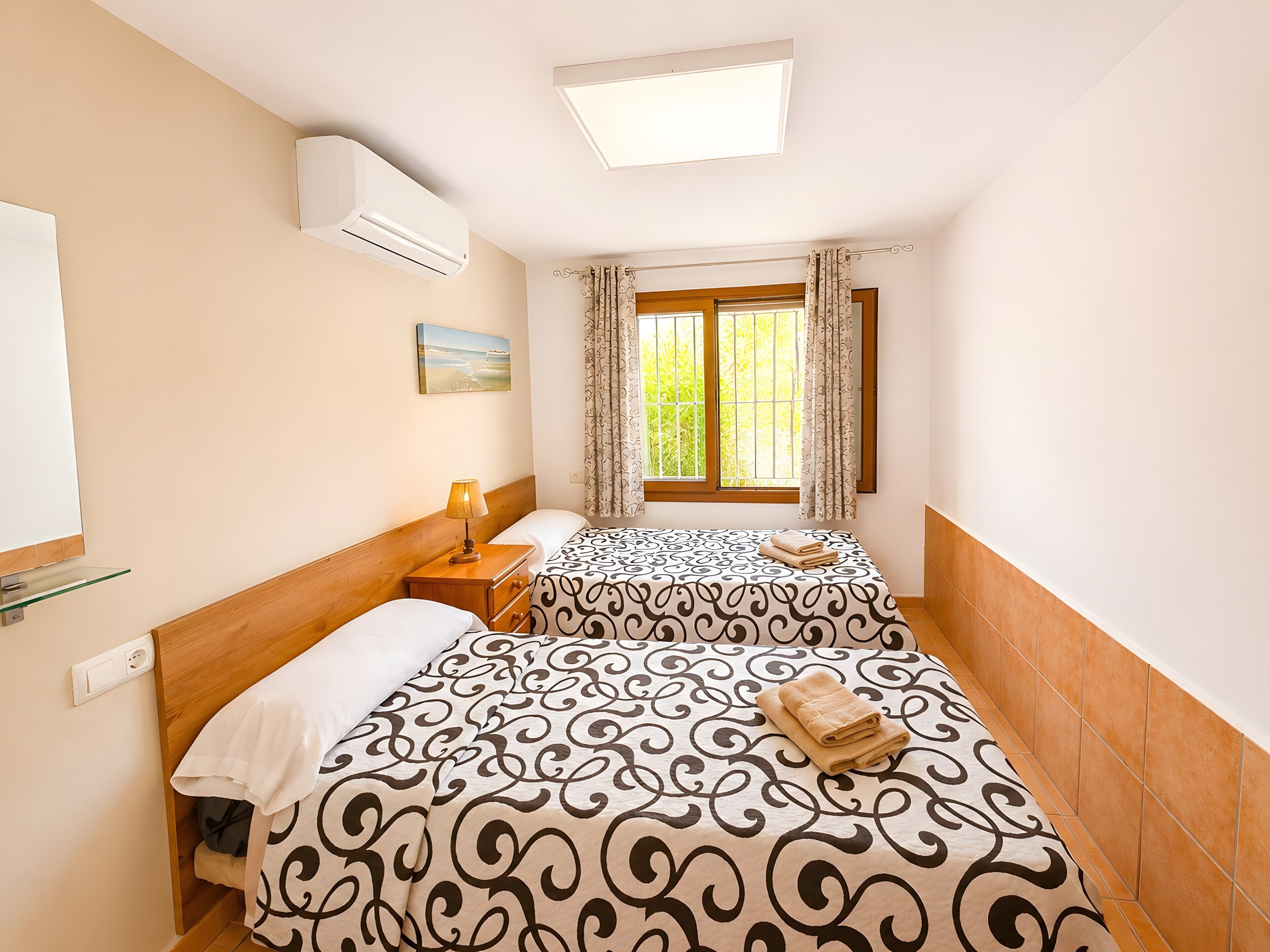 Une chambre d’hôtel avec deux lits, une table de chevet avec une lampe, une fenêtre avec des rideaux et un appareil de climatisation.