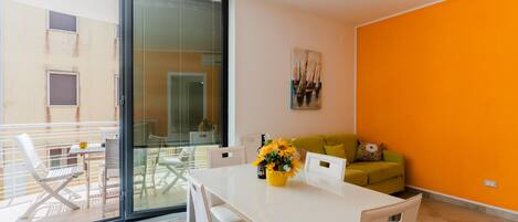 A modern living room with a white table, a yellow vase with flowers, a green sofa, and a large glass door leading to a balcony.