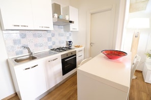 Une cuisine de taille réduite avec des meubles blancs, une cuisinière à gaz, un évier et un plan de travail sur lequel se trouve un saladier rouge.