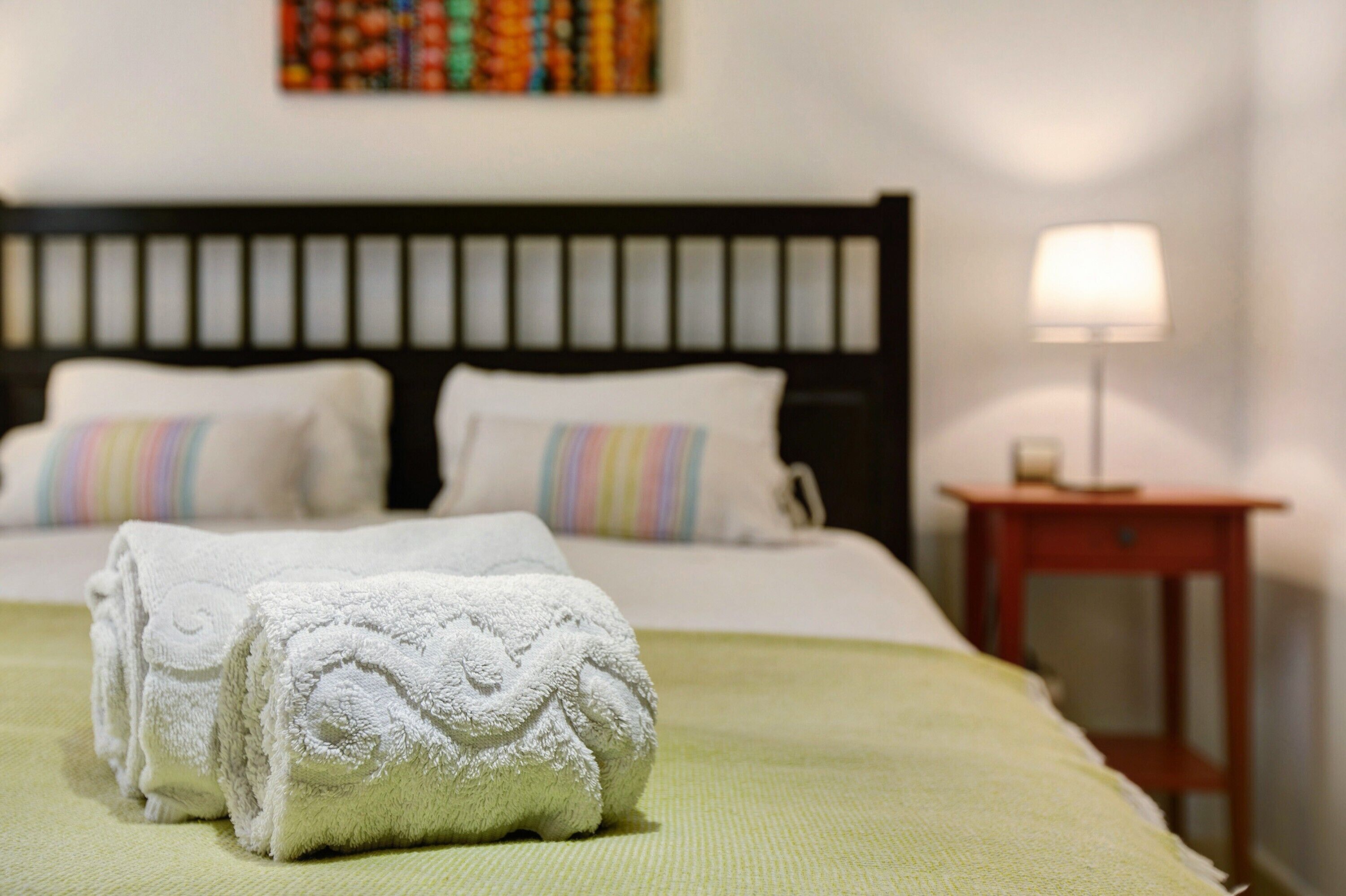 A neatly made bed with white linens and a green bedspread, a bedside table with a lamp, and a colorful abstract painting on the wall.