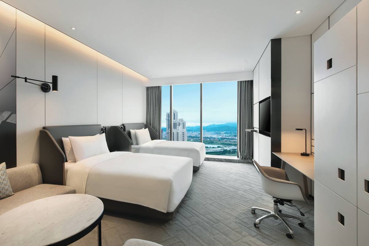 A modern hotel room with two beds, a desk, a chair, and a large window offering a city view.