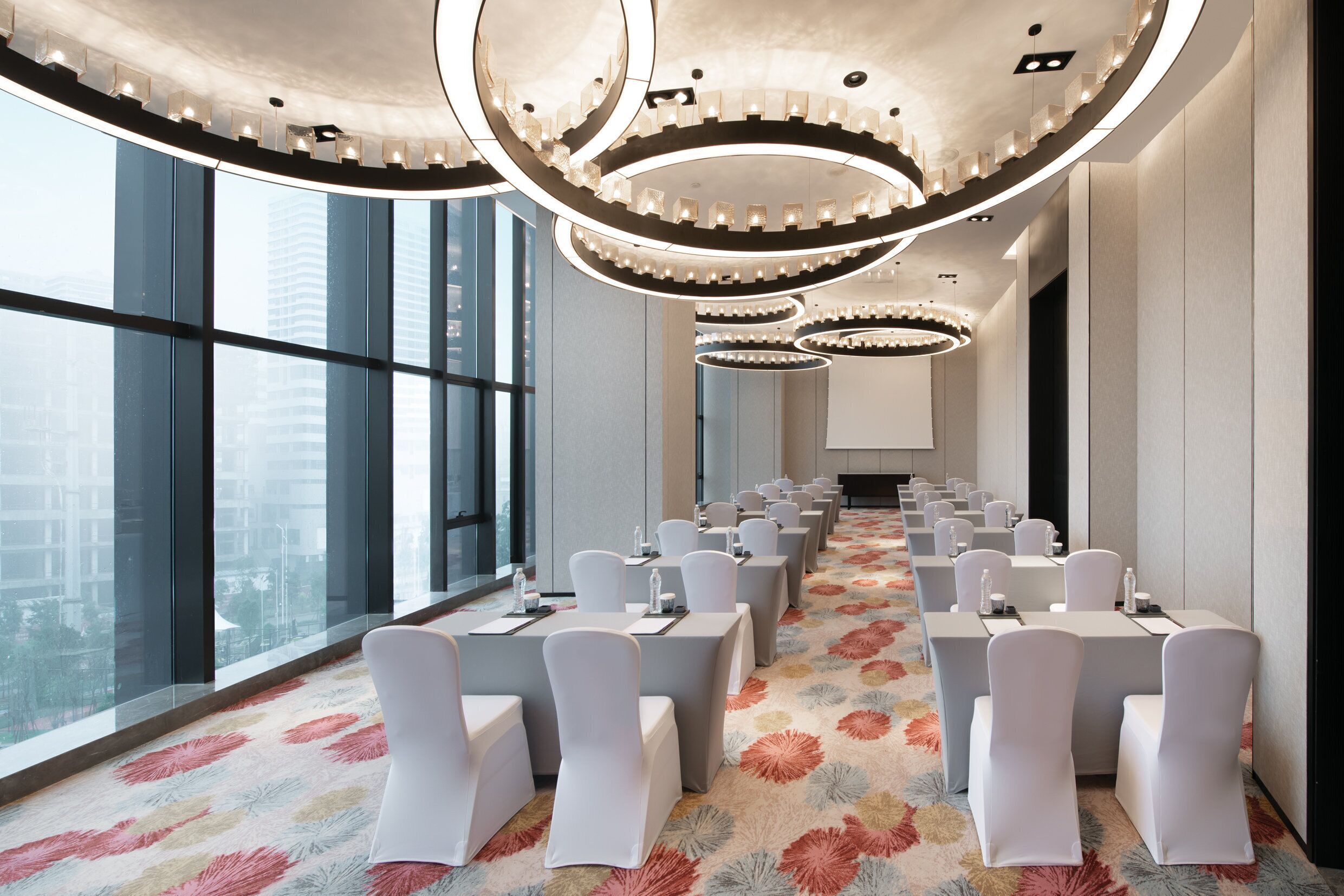 Un salón de conferencias con filas de mesas y sillas, ventanas amplias que ofrecen vistas de la ciudad y un diseño de techo moderno.