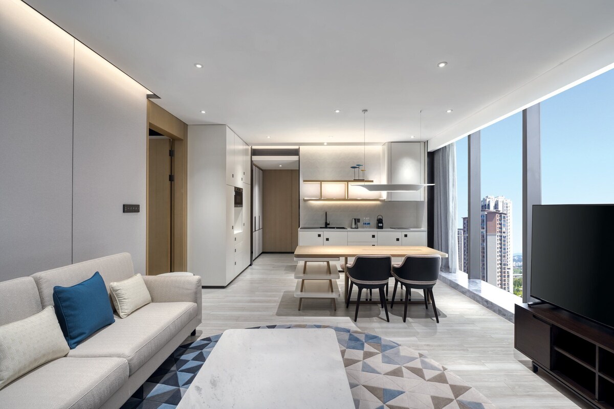 Un salon moderne avec un canapé, une table basse et un téléviseur. Il y a également un coin repas avec une table et des chaises. La pièce est dotée de grandes fenêtres offrant une vue sur la ville.