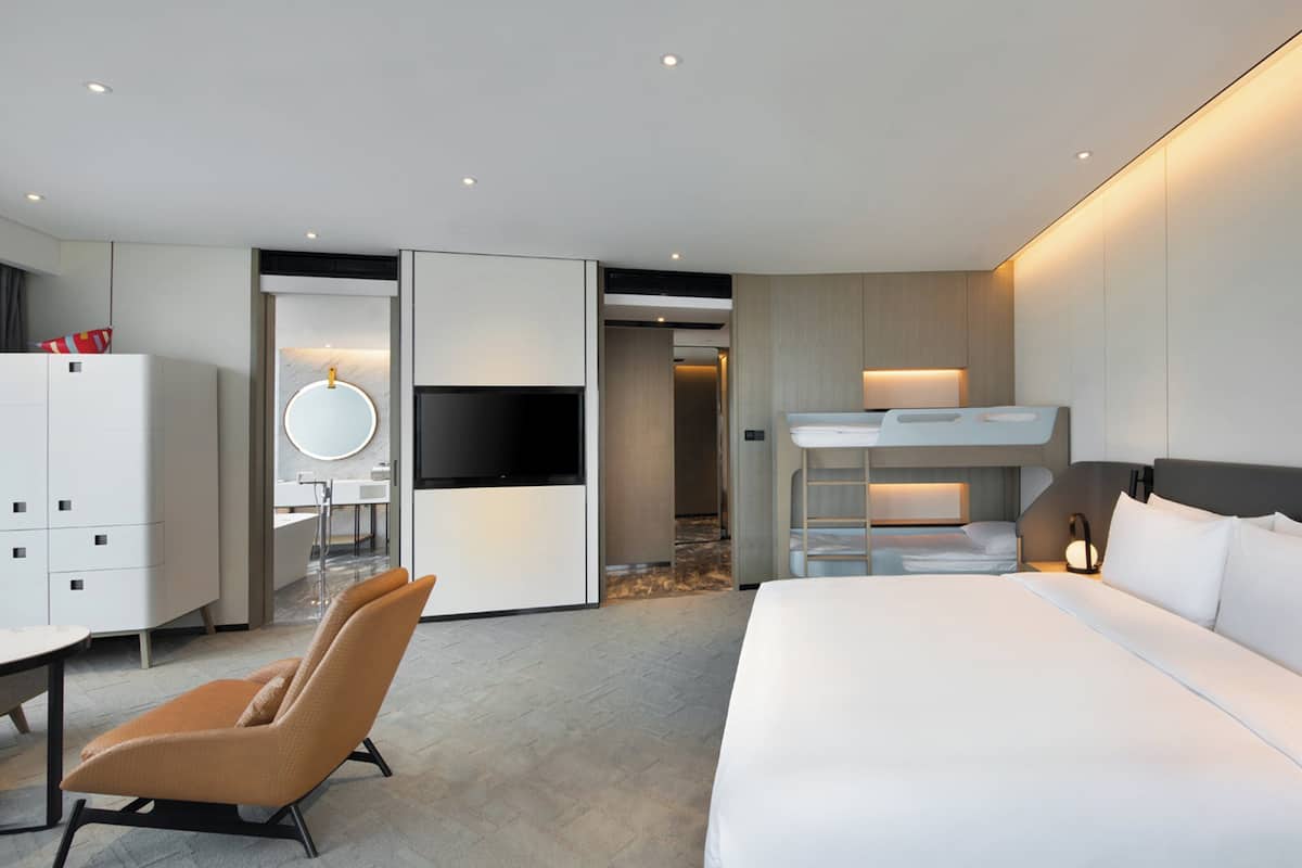A modern hotel room with a large bed, a TV, a desk, and a bunk bed.