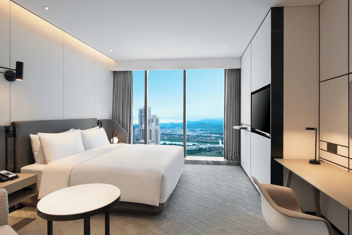 A modern hotel room with a large bed, a desk, a chair, a TV, and a view of the cityscape.