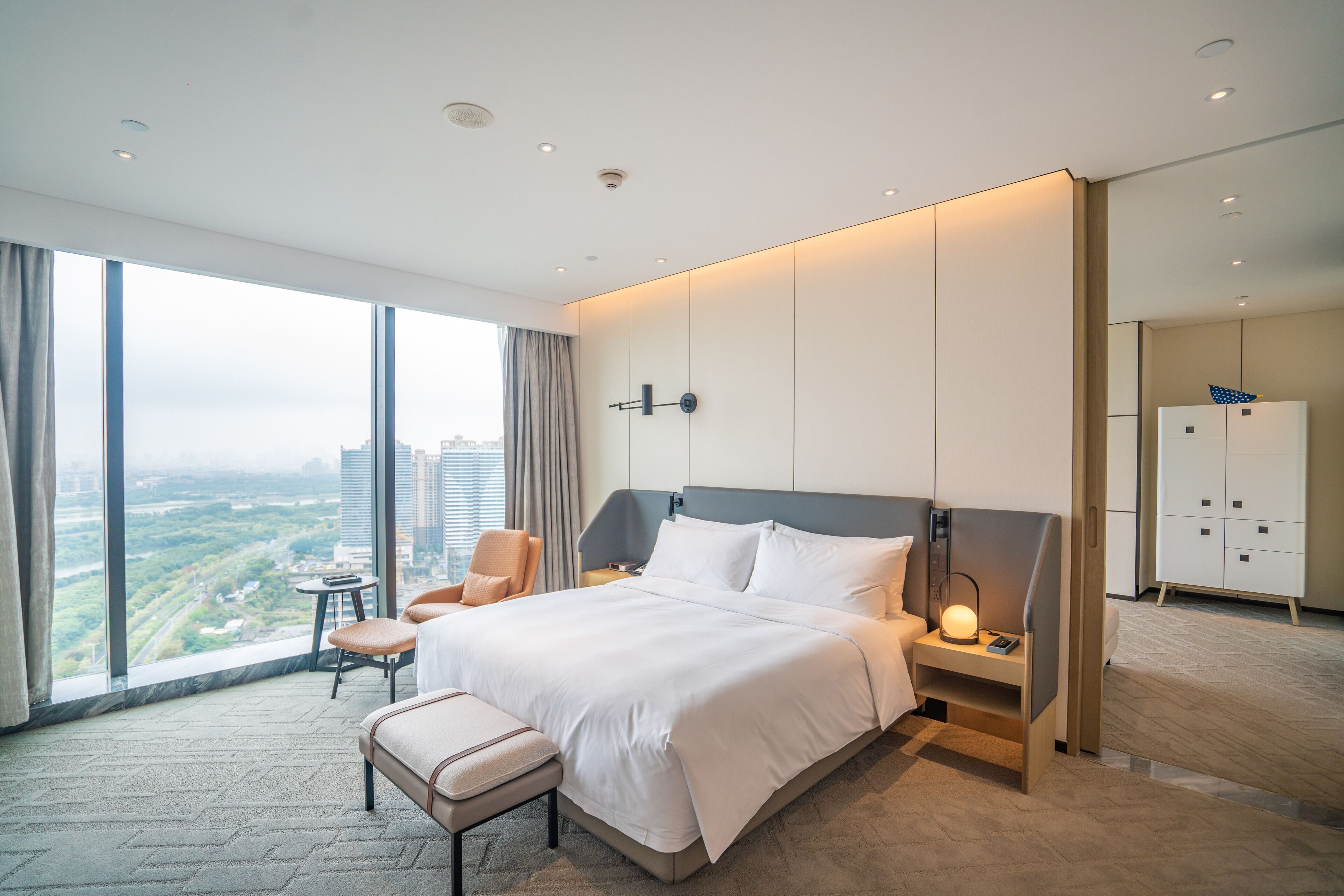 A modern hotel room with a large bed, a bedside table with a lamp, a chair, and a view of the city.