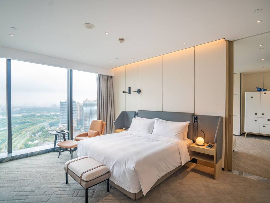 A modern hotel room with a large bed, a bedside table with a lamp, a chair, and a view of the city.