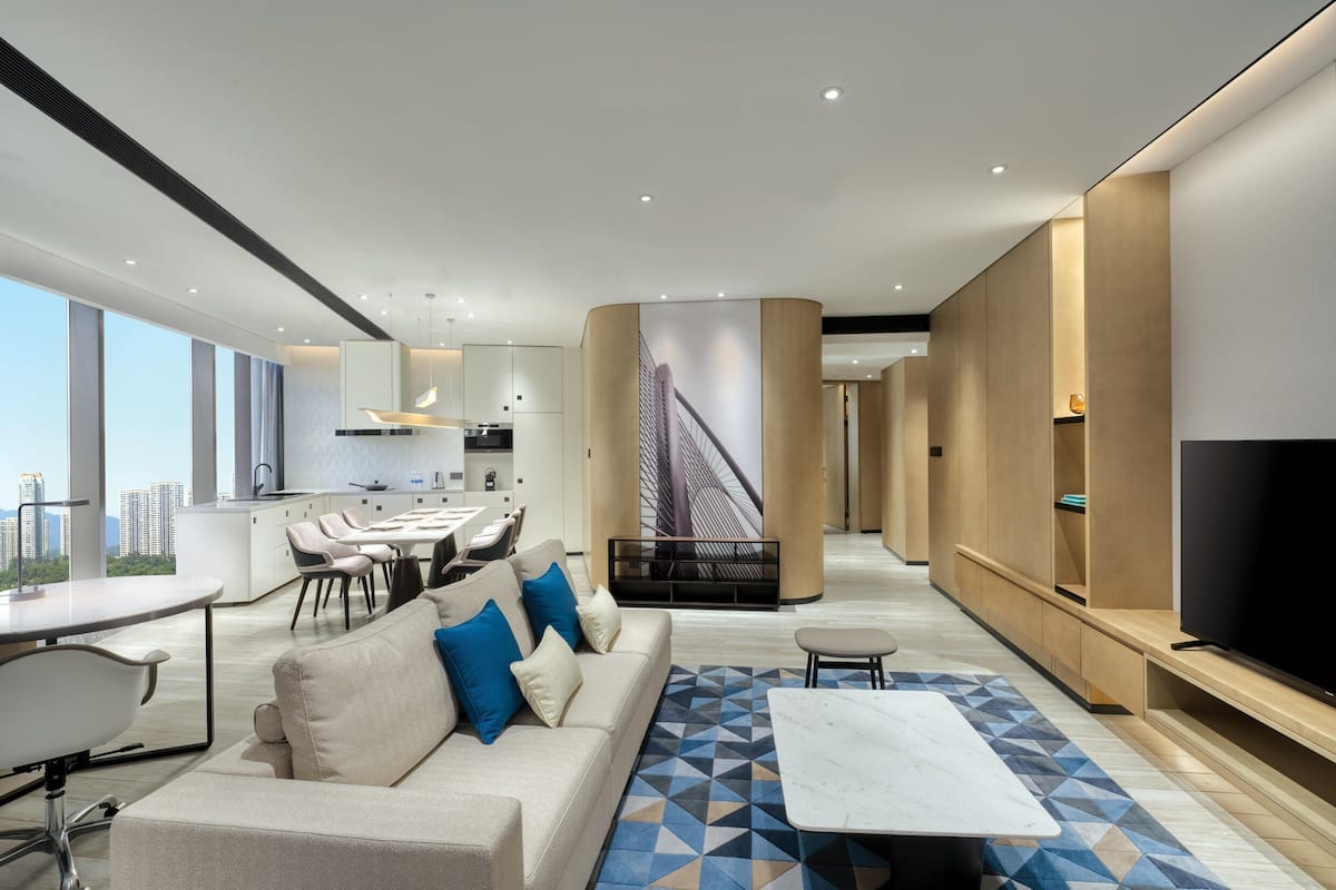A modern living room with a sofa, coffee table, and TV. There is a dining area with chairs and a table. The room has large windows and a view of the city.