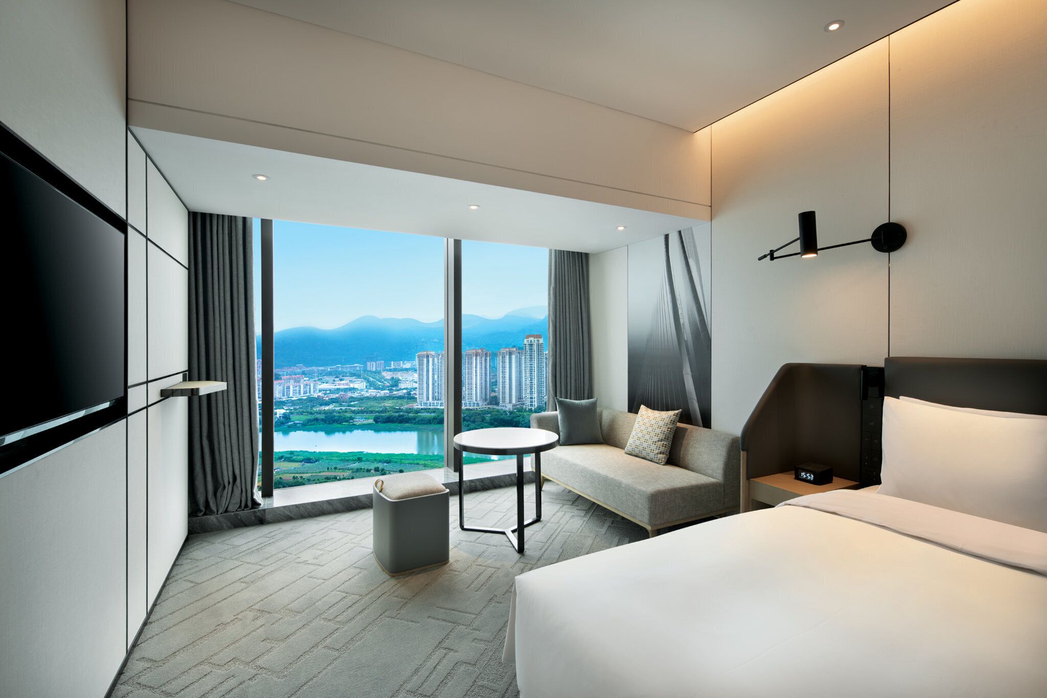 A modern hotel room with a large window offering a city view, a bed, a sofa, a small table, and a TV.