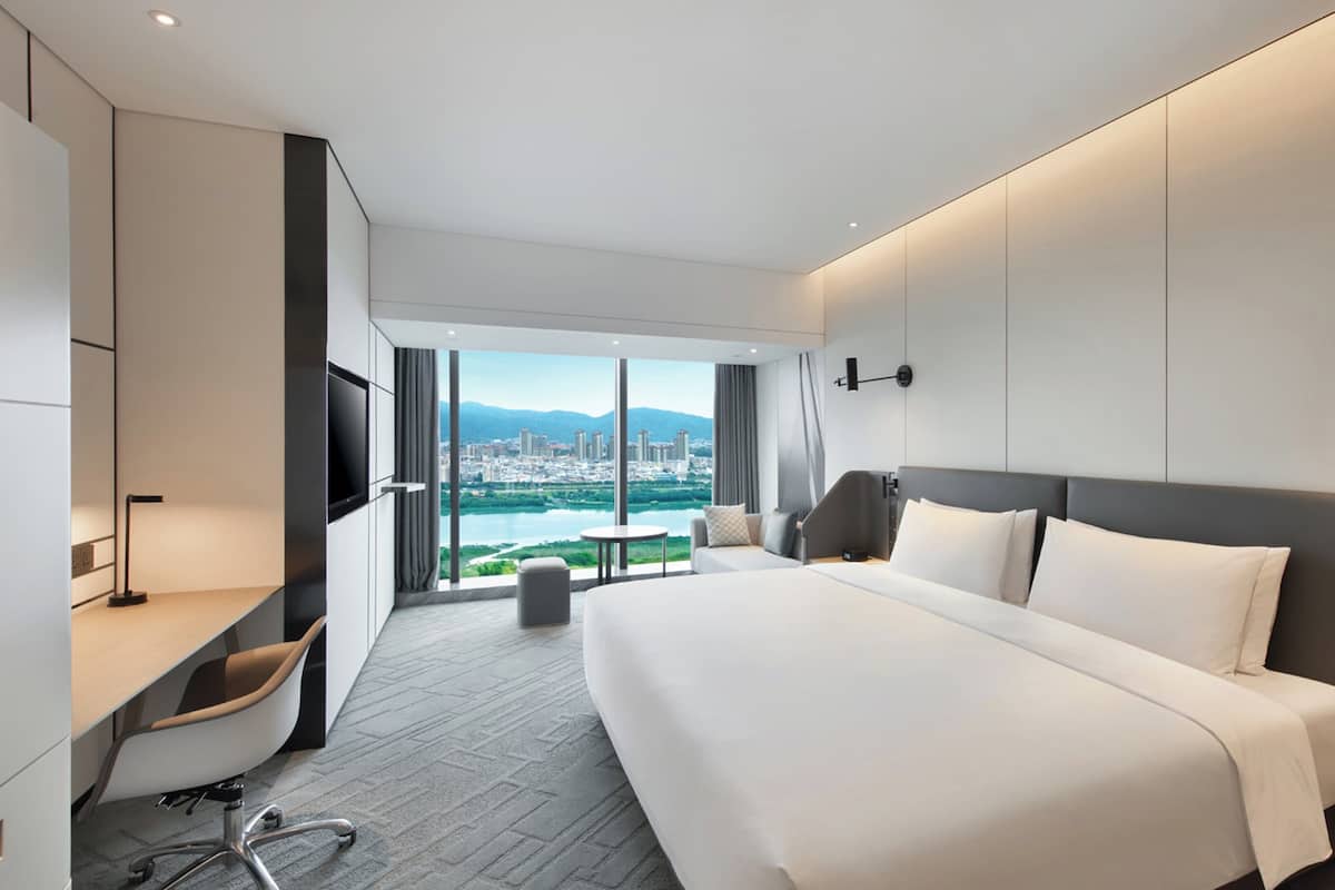 A modern hotel room with a large bed, a desk, a chair, a TV, and a view of a cityscape.