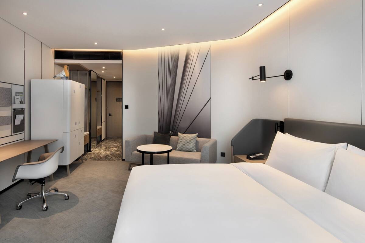 A modern hotel room with a large bed, a desk with a chair, a small round table, and a sofa.