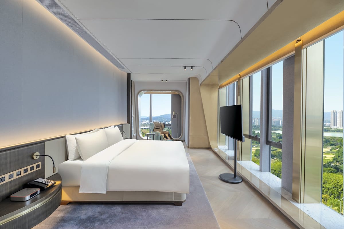 A modern hotel room with a large bed, a TV, and a city view through large windows.