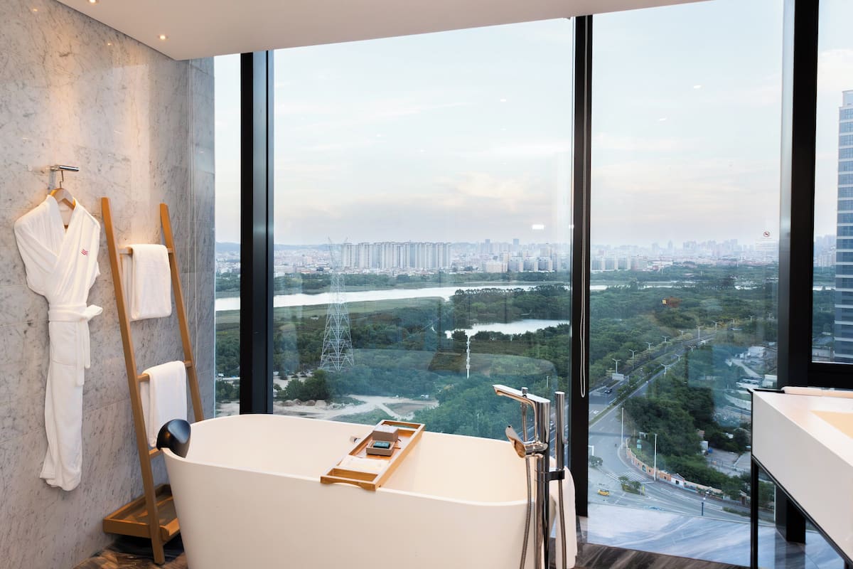 A modern bathroom with a large bathtub, a wooden towel rack, and a view of a cityscape through large windows.