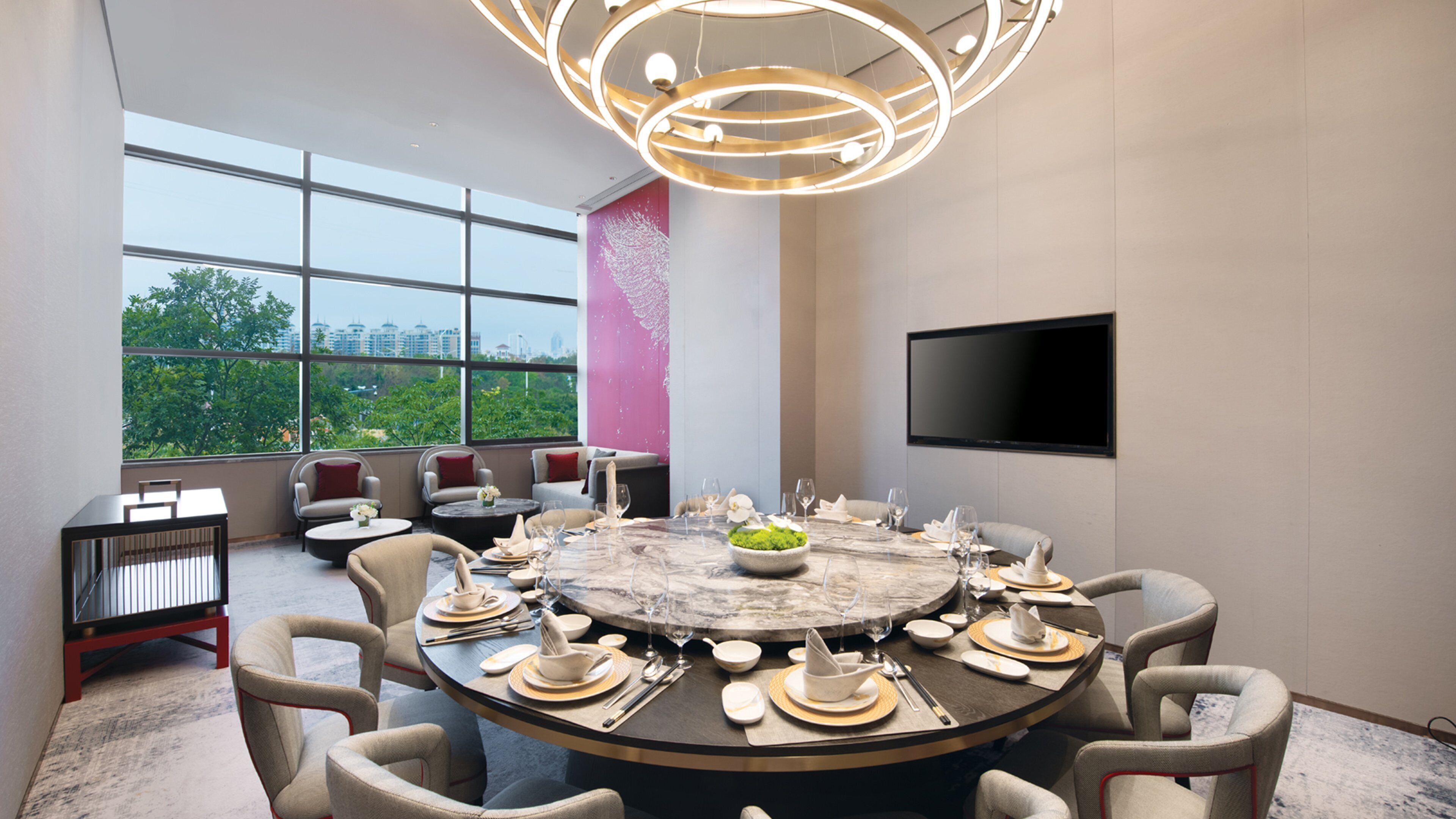 A modern conference room with a large round table set for dining, surrounded by comfortable chairs, a large window offering a city view, and a mounted flat-screen TV on the wall.