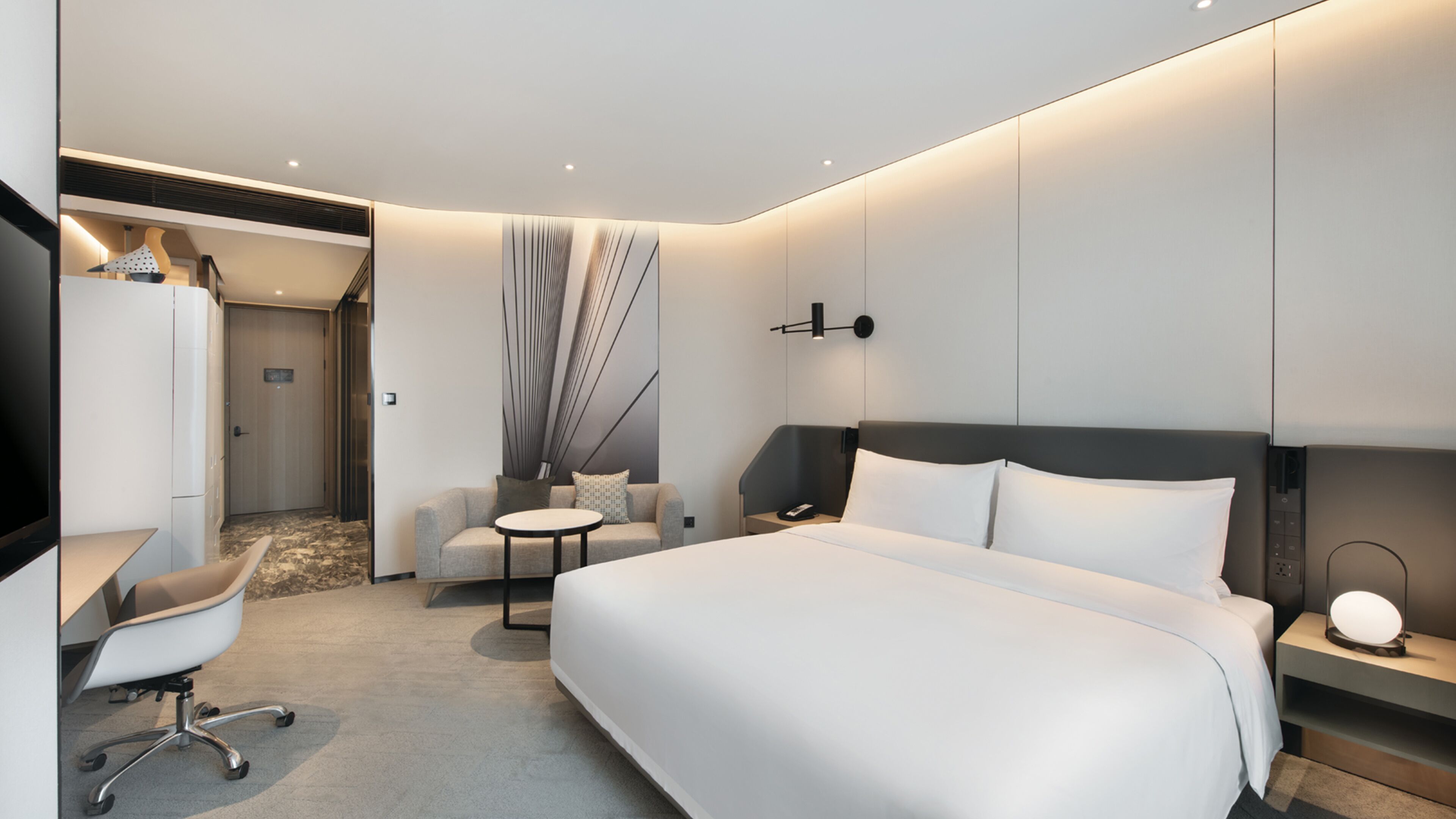 A modern hotel room with a large bed, a desk with a chair, a small round table, and a sofa.