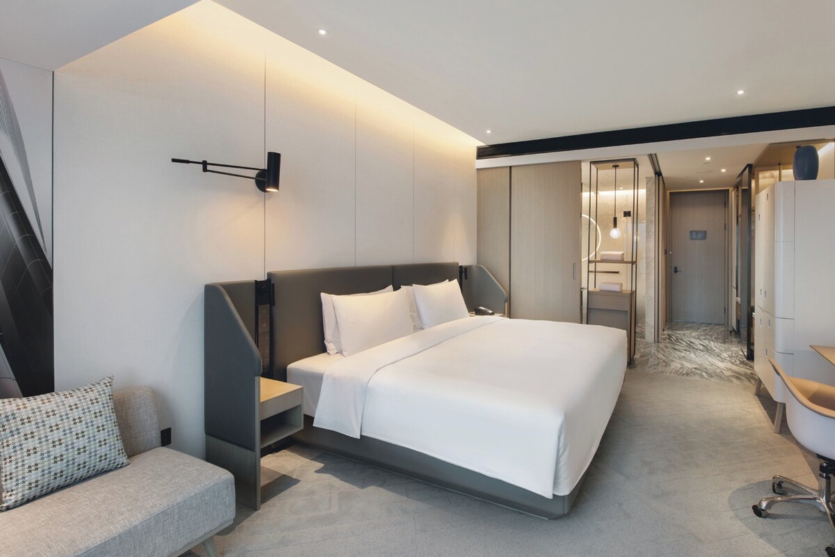 Une chambre d’hôtel moderne dotée d’un grand lit, d’un canapé, d’une télévision et d’une salle de bain visible à travers une porte en verre.