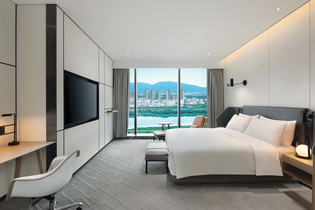 Une chambre d’hôtel moderne dotée d’un grand lit, d’un bureau, d’une télévision et offrant une vue sur le paysage urbain grâce à de grandes fenêtres.