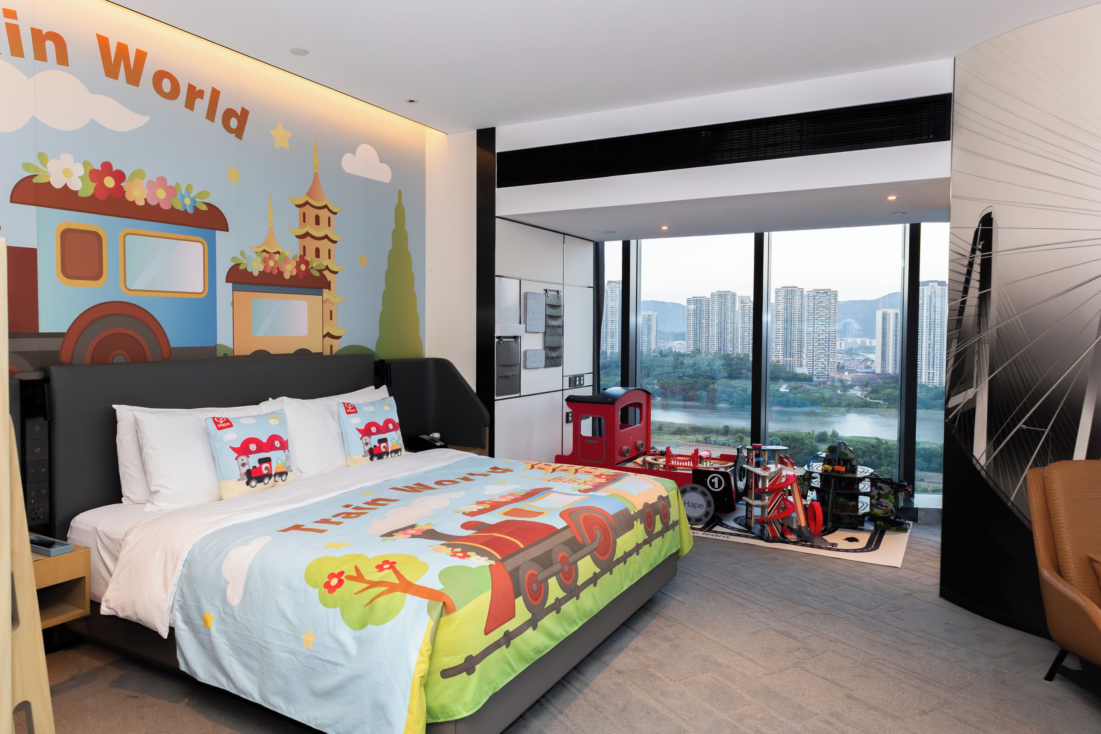 A modern bedroom with a large bed, a city view, and a train-themed bedspread.