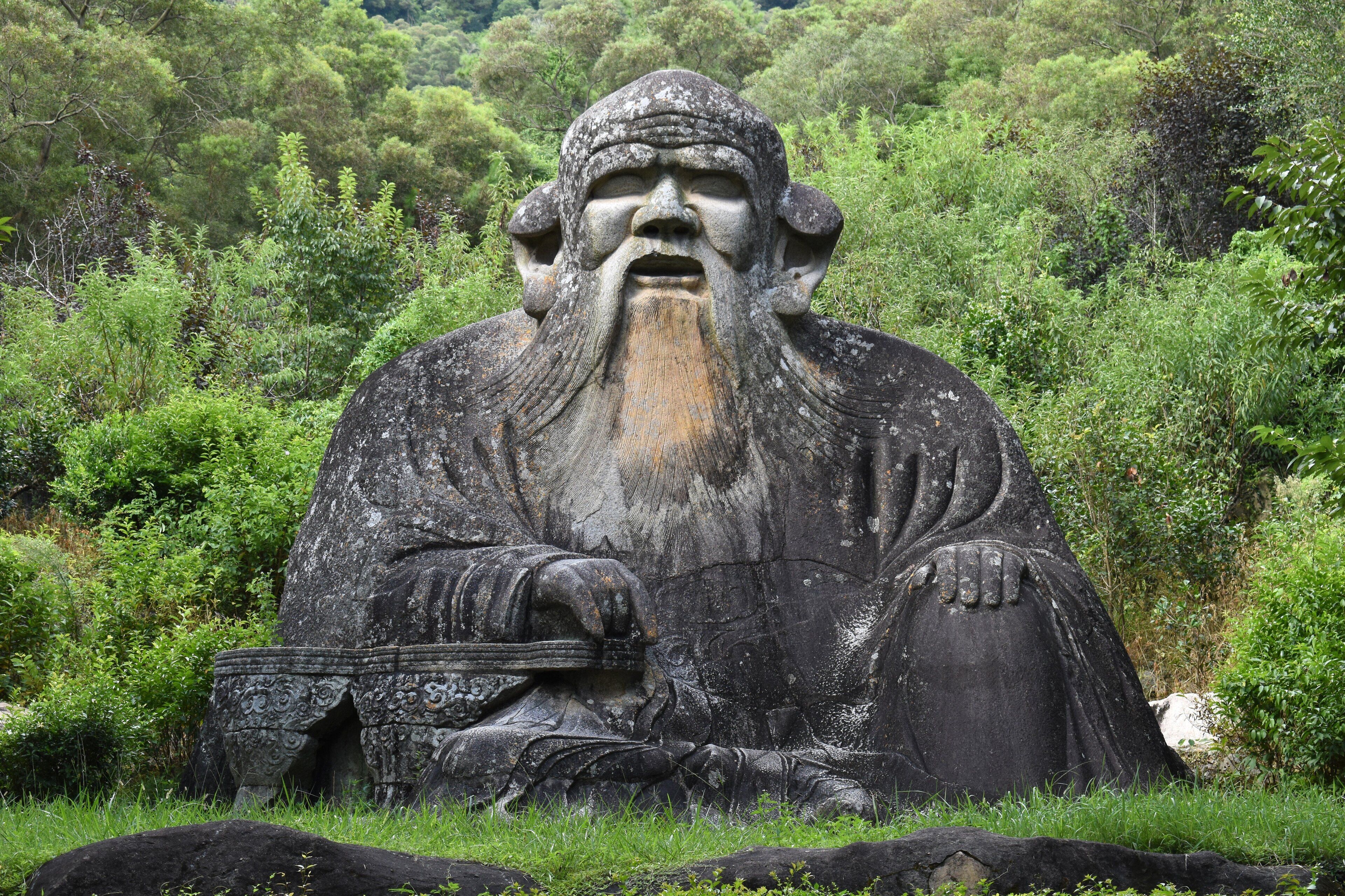 Una gran estatua de piedra de una figura sentada con una barba larga, rodeada de vegetación exuberante.