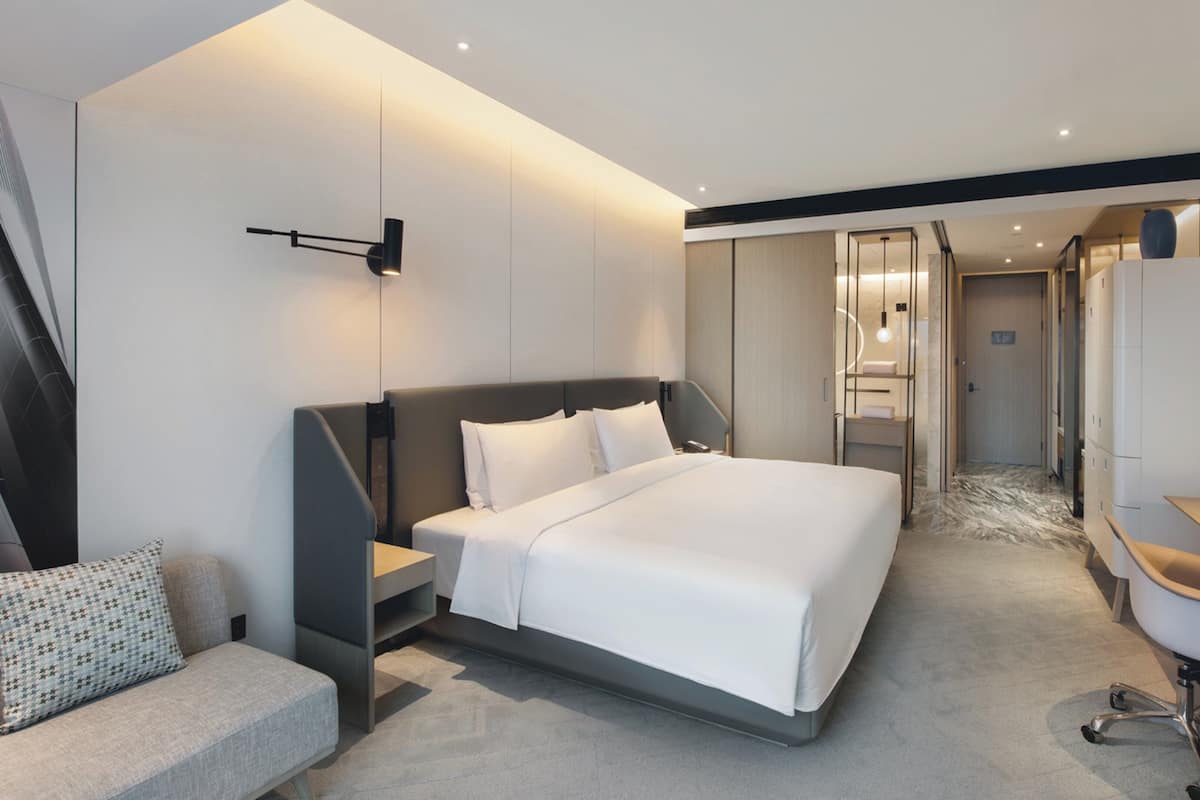 A modern hotel room with a large bed, a sofa, a TV, and a bathroom visible through a glass door.