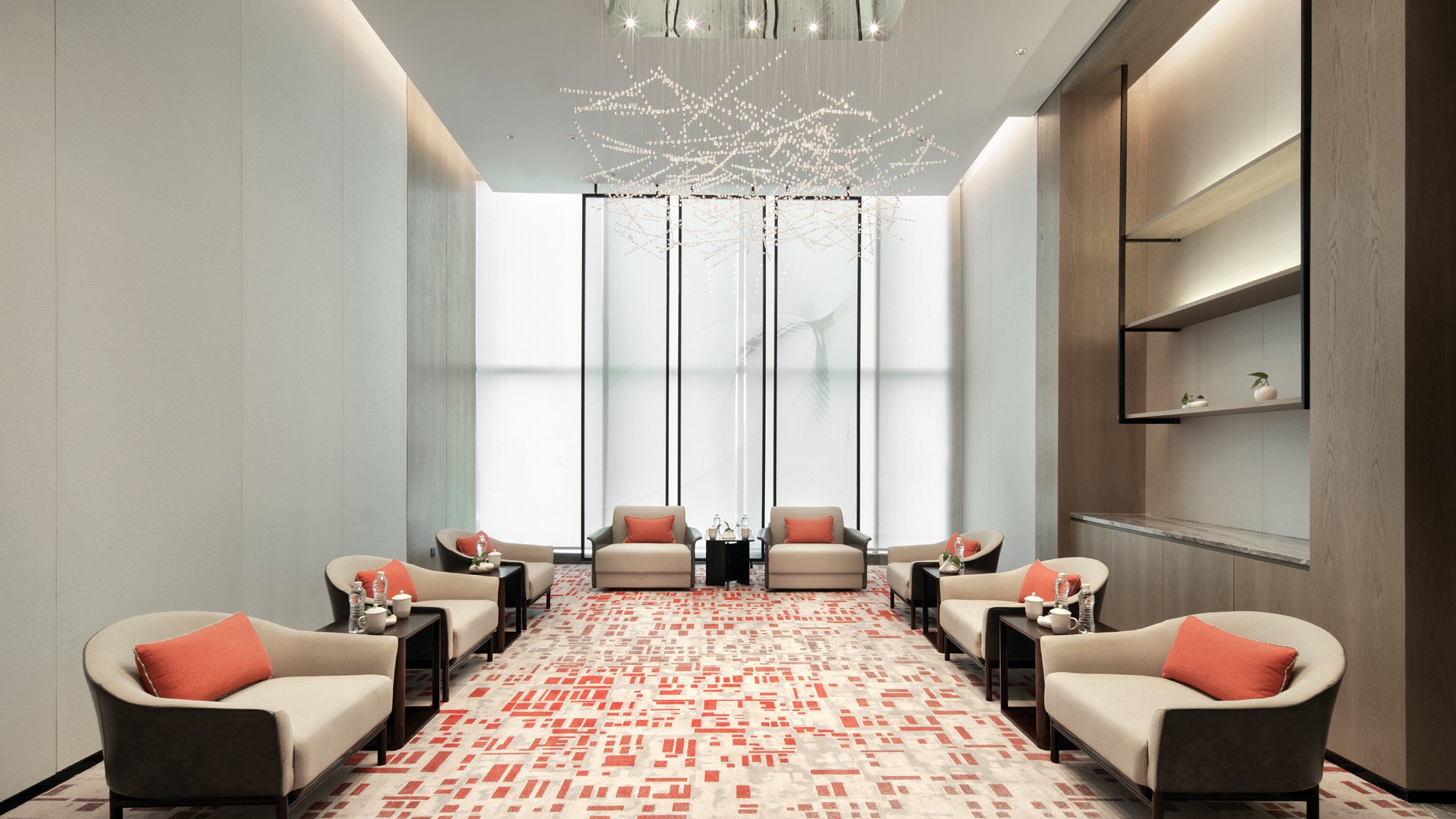 A modern, well-lit room with a patterned carpet, seating area with armchairs and side tables, a large window with vertical blinds, and a decorative ceiling light fixture.