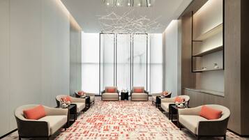 A modern, well-lit room with a patterned carpet, seating area with armchairs and side tables, a large window with vertical blinds, and a decorative ceiling light fixture.